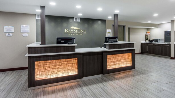 Uma mesa de recepção com parede escura e painéis iluminados, um balcão e uma placa com a inscrição 'Baymont Inn & Suites'.