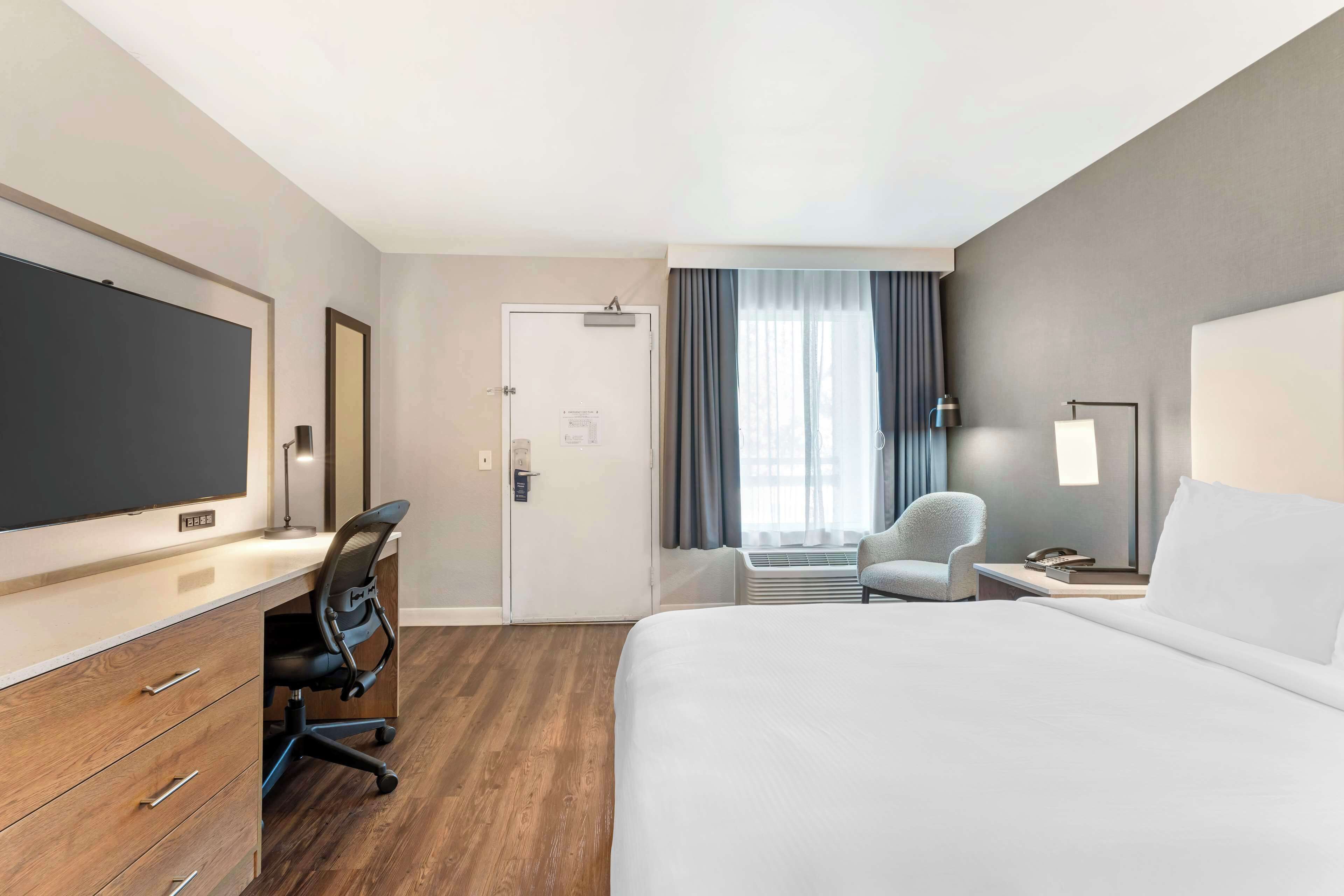 A hotel room with a bed, a desk with a chair, a television, a door, and a window with curtains.