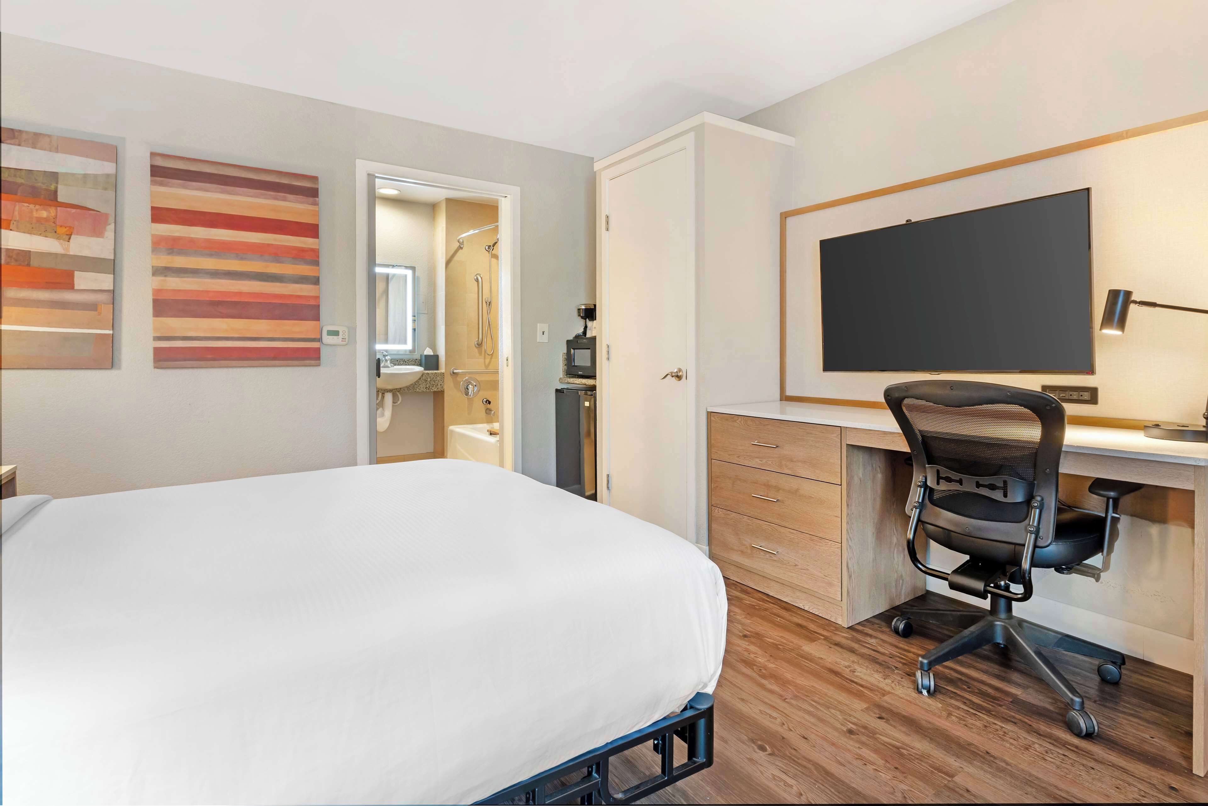 A hotel room with a bed, a desk with a computer, a TV, and artwork on the walls.