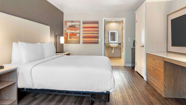 A modern hotel room with a large bed, a bedside table, a wardrobe, and a bathroom visible through an open door.