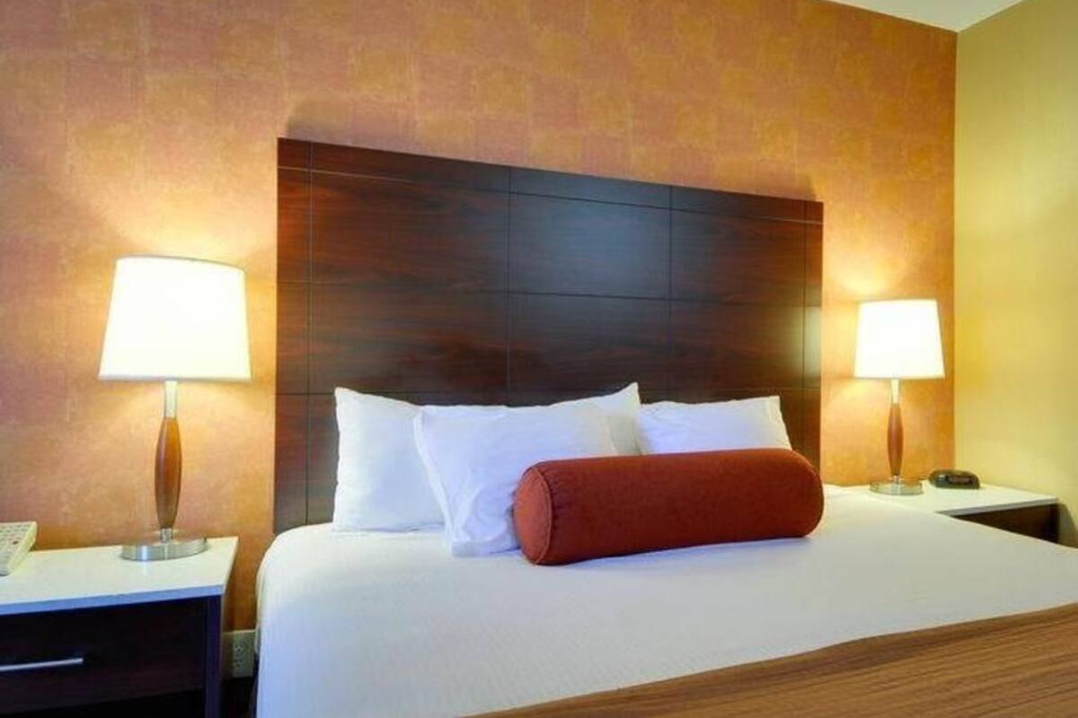 A hotel room with a large bed, two bedside lamps, a wooden headboard, and a red accent pillow.