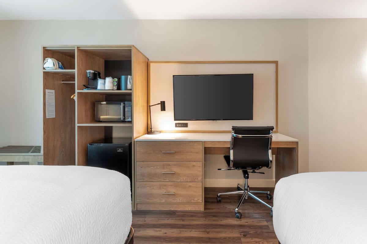 A hotel room with two beds, a wooden desk with a computer, a flat-screen TV, and a kitchenette with a microwave and coffee maker.