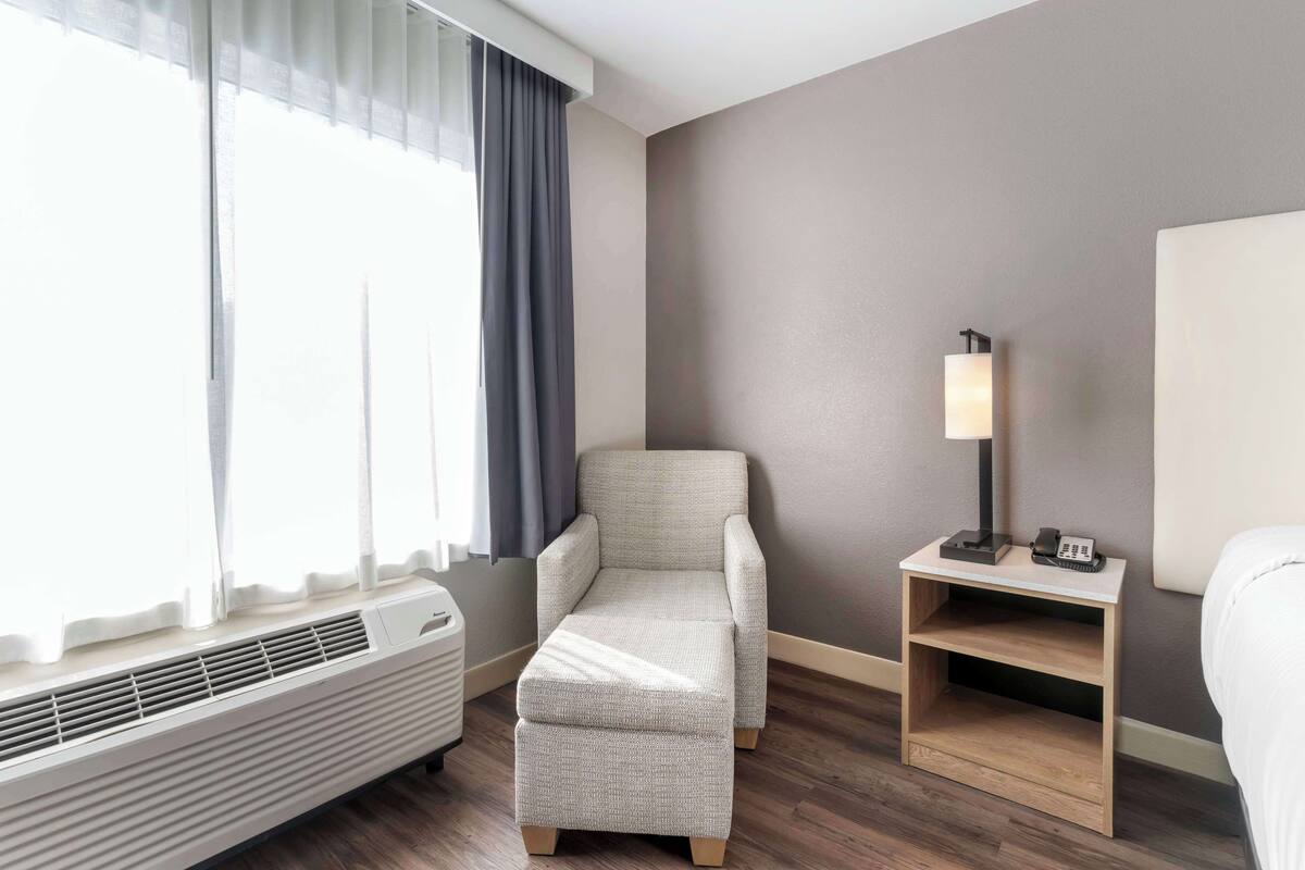 A hotel room with a bed, a chair, a nightstand with a lamp, and a heater.