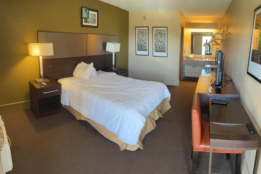 Ein Hotelzimmer mit Bett, Nachttischen, Schreibtisch mit Stuhl, Fernseher und einem Badezimmer mit Spiegel und Waschbecken.