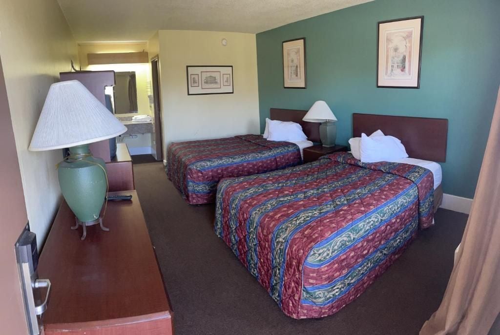 A hotel room with two beds, a desk, a lamp, and a mirror.