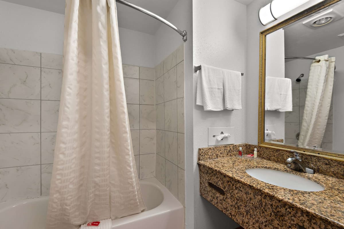 A bathroom with a bathtub, a sink with a granite countertop, a mirror with a gold frame, and white towels on the wall.