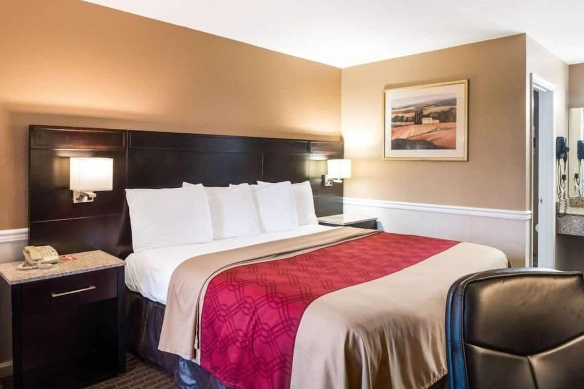A hotel room with a bed, bedside lamps, a telephone, a chair, and a painting on the wall.