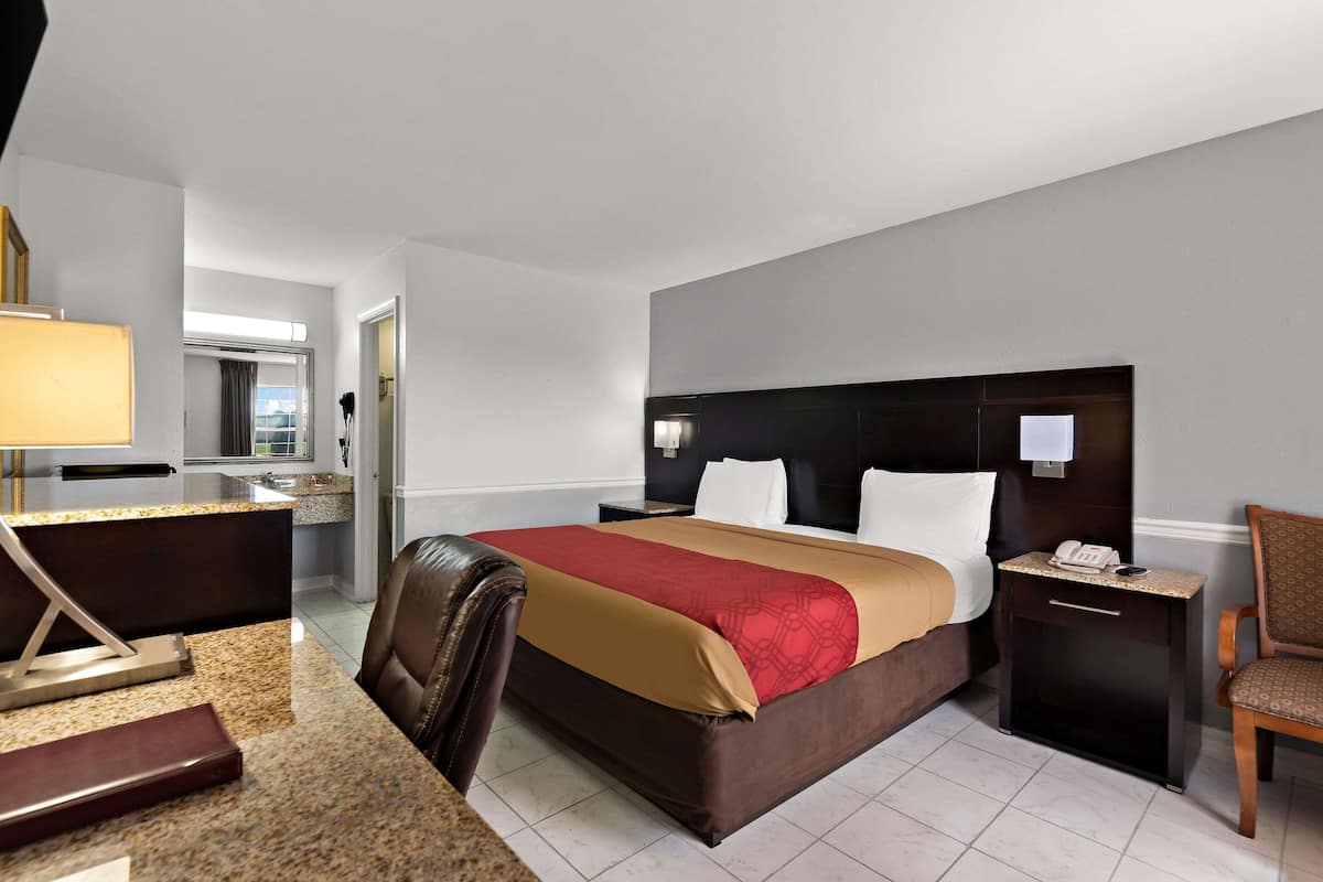 A hotel room with a bed, a desk with a lamp, a chair, a nightstand, and a small table.