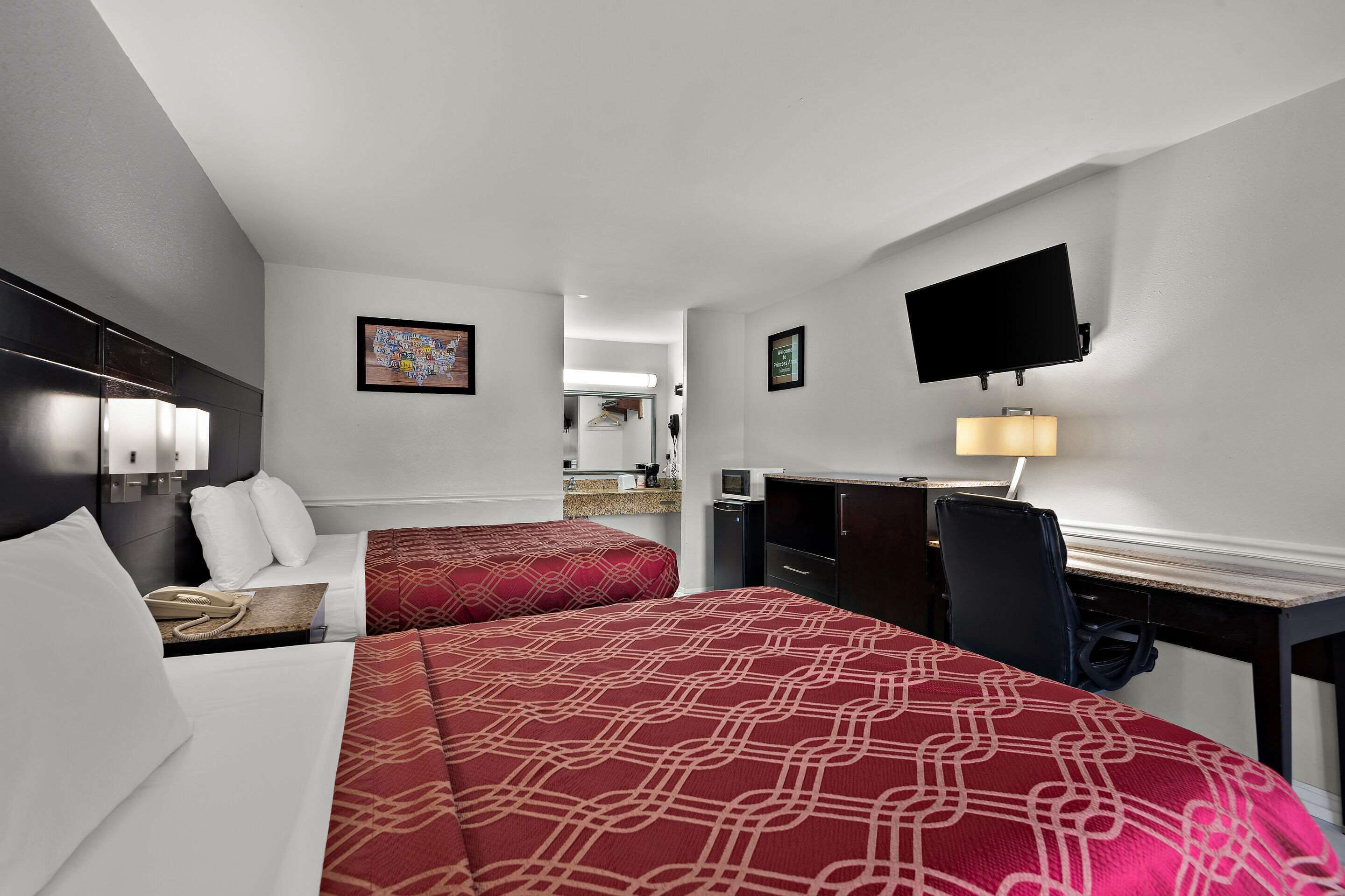 A hotel room with two beds, a desk, a chair, a TV mounted on the wall, and a lamp.