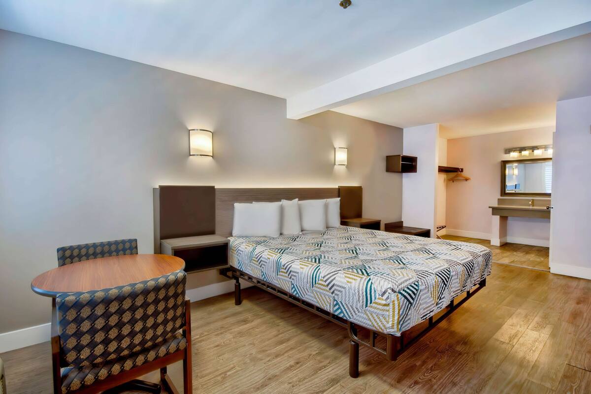 A hotel room with a bed, a small table with a chair, a bedside table, a mirror, and a wall-mounted TV.