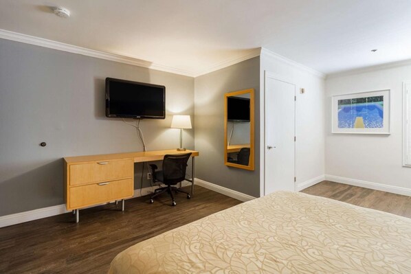 Une chambre d’hôtel comprenant un lit, un bureau avec une chaise, un téléviseur à écran plat fixé au mur, un miroir et un tableau encadré accroché au mur.