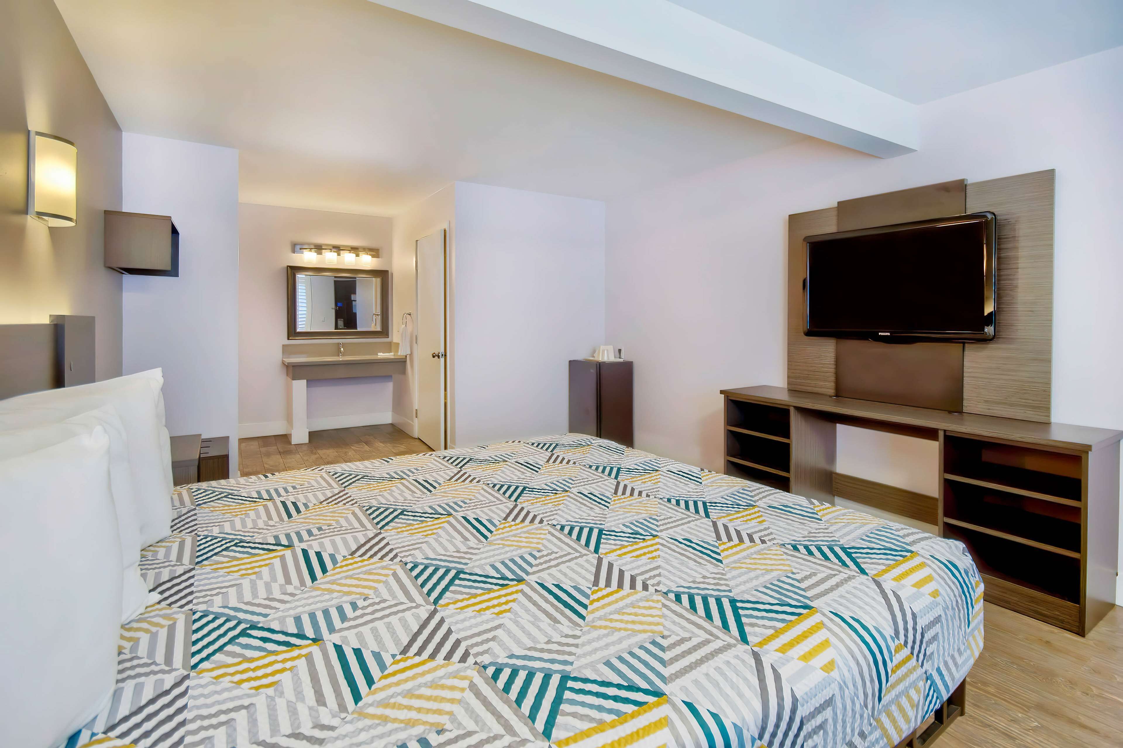 A hotel room with a bed, a TV mounted on the wall, a vanity with a mirror, and a small seating area.