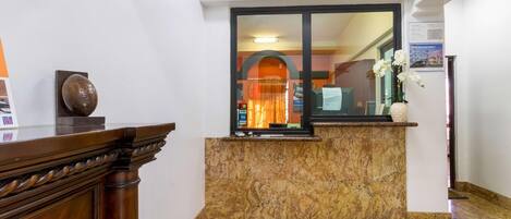 Uma área de recepção com piso de mármore, balcão de madeira, janela com vista para o interior e um vaso com flores sobre o balcão.