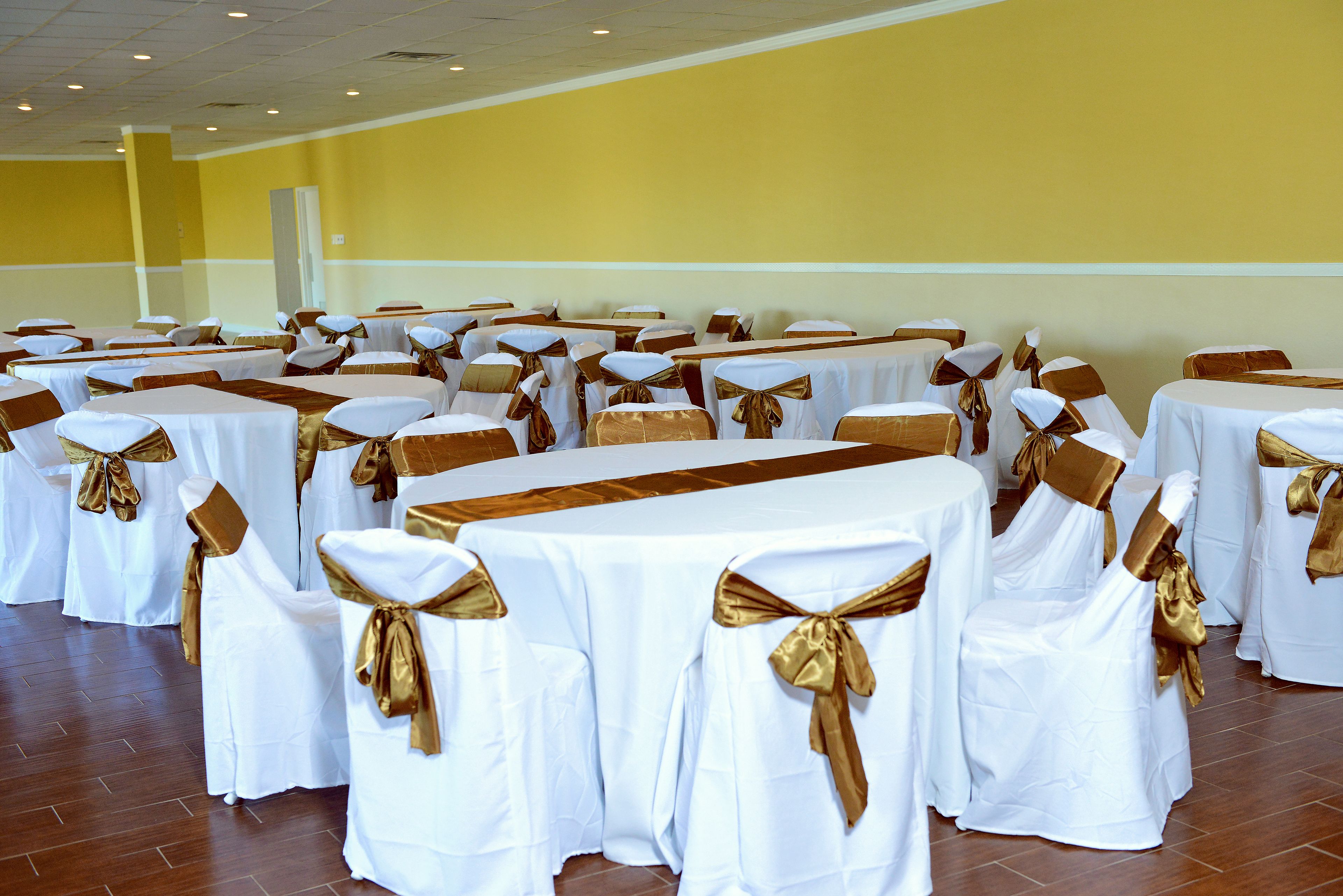 Une salle de banquet avec des rangées de tables rondes, chacune recouverte de nappes blanches et ornée de rubans dorés, et des chaises assorties, également dotées de rubans dorés.