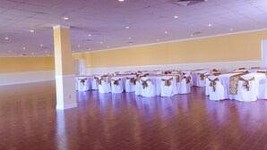 Une grande salle vide, dotée d’un plancher en bois et de tables blanches ornées de rubans dorés, probablement un espace destiné à des banquets ou à l’organisation d’événements.