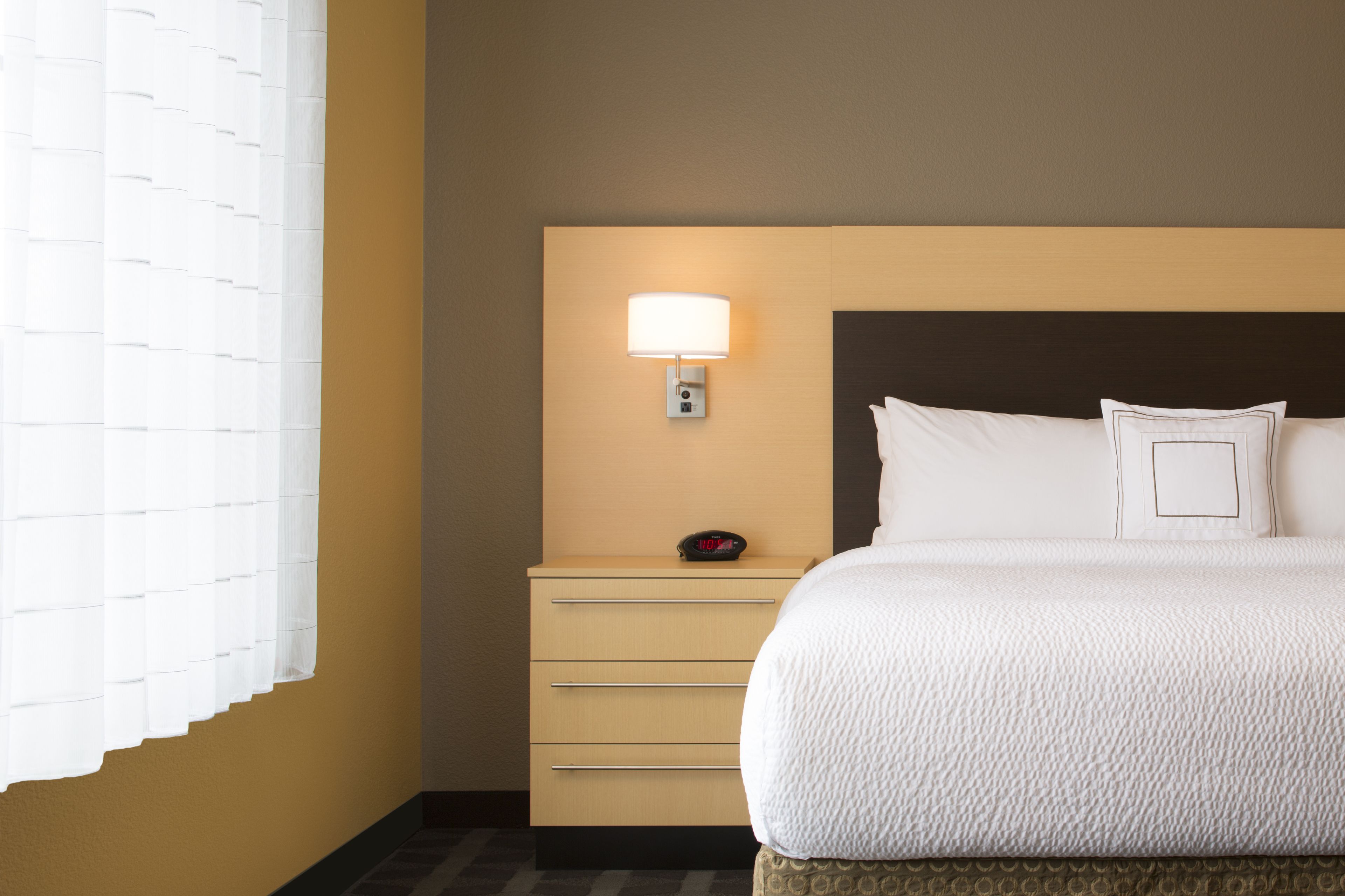 A hotel room with a bed, a nightstand, a wall-mounted lamp, and a window with blinds.