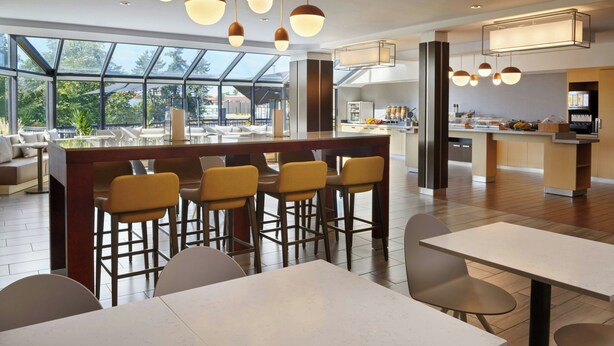 A modern restaurant with a bar area, high ceilings, and large windows offering views of the outdoors. - TownePlace Suites by Marriott London (London)