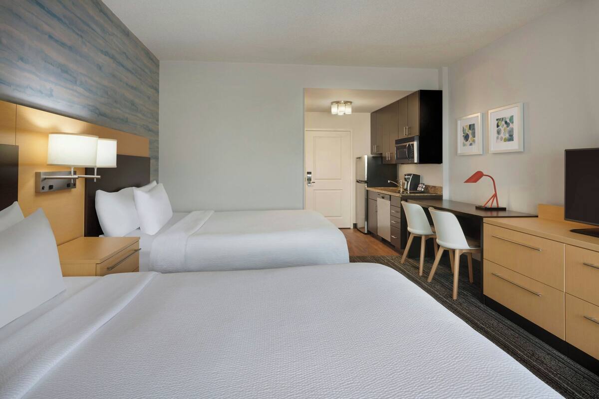 A hotel room with two beds, a desk, a chair, a TV, and a kitchenette.