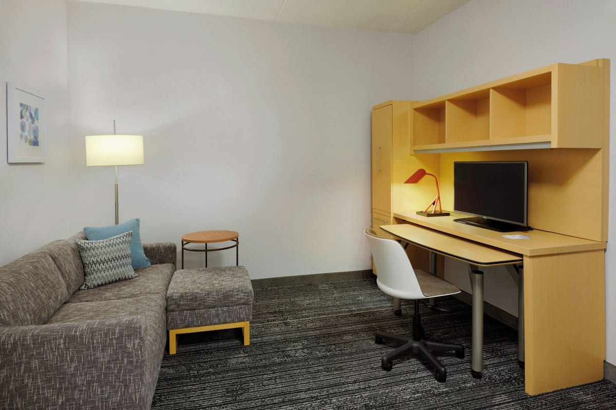 A modern hotel room with a grey sofa, a small round table, a desk with a computer and lamp, and a TV on a wooden shelf.