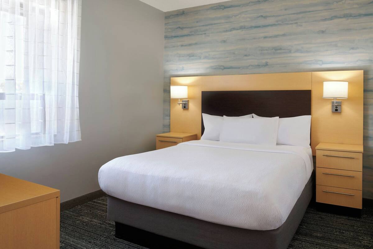 A hotel room with a large bed, two bedside tables, a wooden nightstand, and a window with sheer curtains.