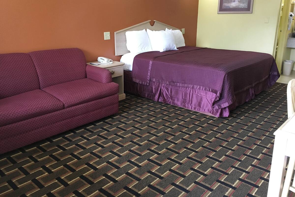 Hotellihuone, jossa on violetti sohva, violetilla päiväpeitteellä varustettu sänky, yöpöytä, jossa on puhelin, ja kuvioitu matto.