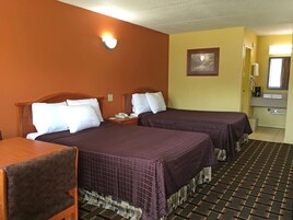 Quarto de hotel com duas camas, cabeceira de madeira, mesa de cabeceira com telefone, pequena mesa, uma cadeira, banheiro com pia e espelho, e um quadro na parede.