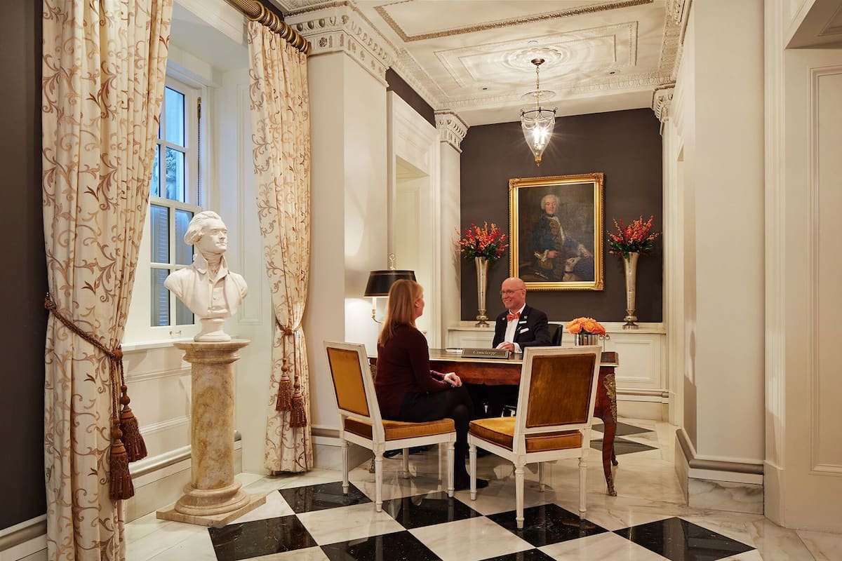 A formal room with a checkered floor, a bust on a pedestal, a desk with two chairs, and a painting on the wall.