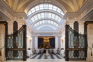 An opulent room with a high arched skylight, a checkered floor, and a formal dining setup with a floral centerpiece.