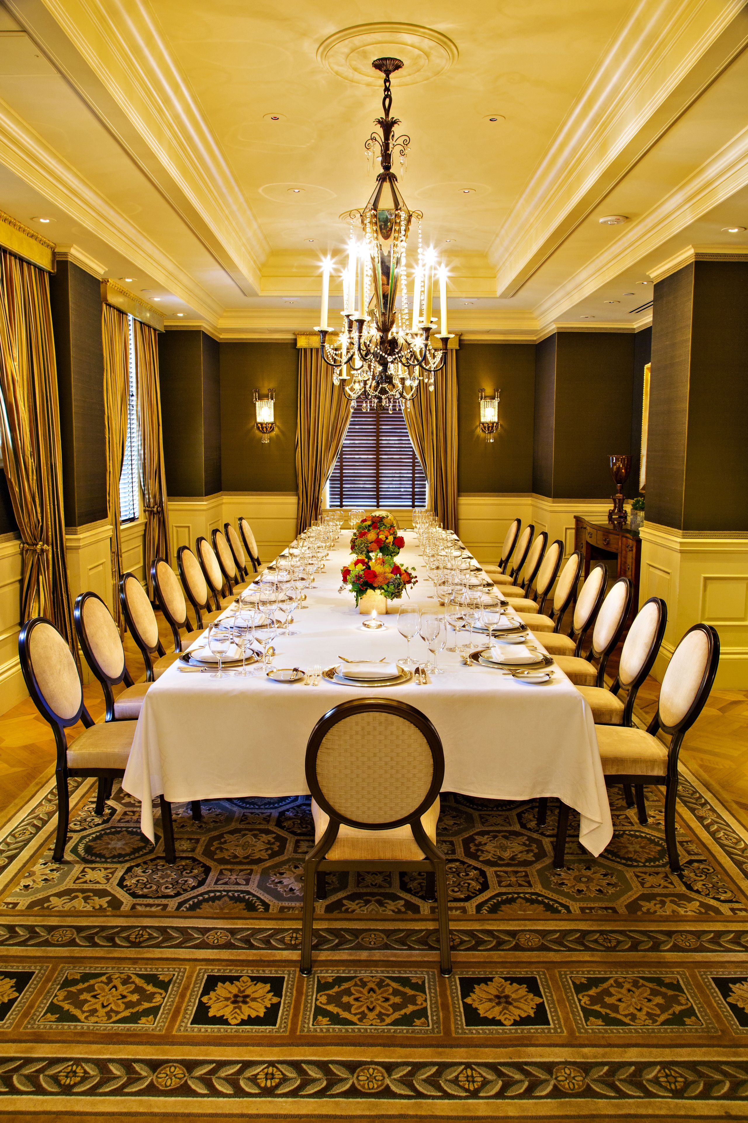 A formal dining room with a long table set for a meal, flanked by chairs, a chandelier, and patterned carpet.