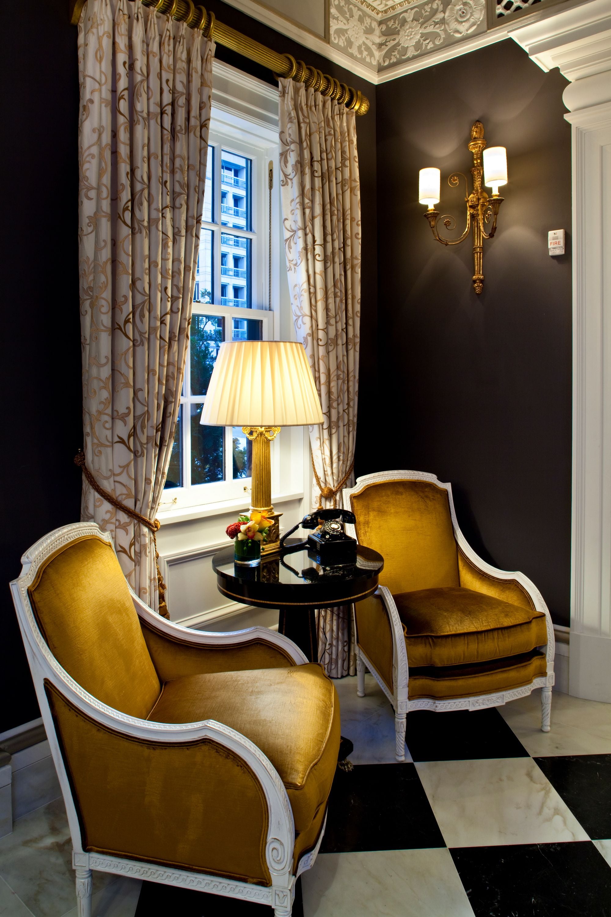 Een kamer met twee gouden fauteuils, een tafel met een lamp en een raam met gordijnen.