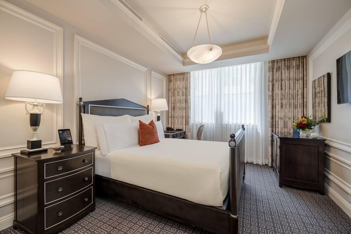 A hotel room with a large bed, two bedside tables with lamps, a dresser, a large window with curtains, and a wall-mounted TV.