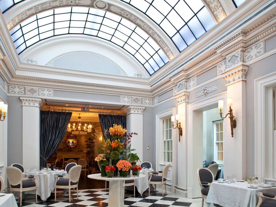 Un comedor amplio y elegante con techo alto y tragaluces, que presenta columnas ornamentadas, candelabros y suelo a cuadros.