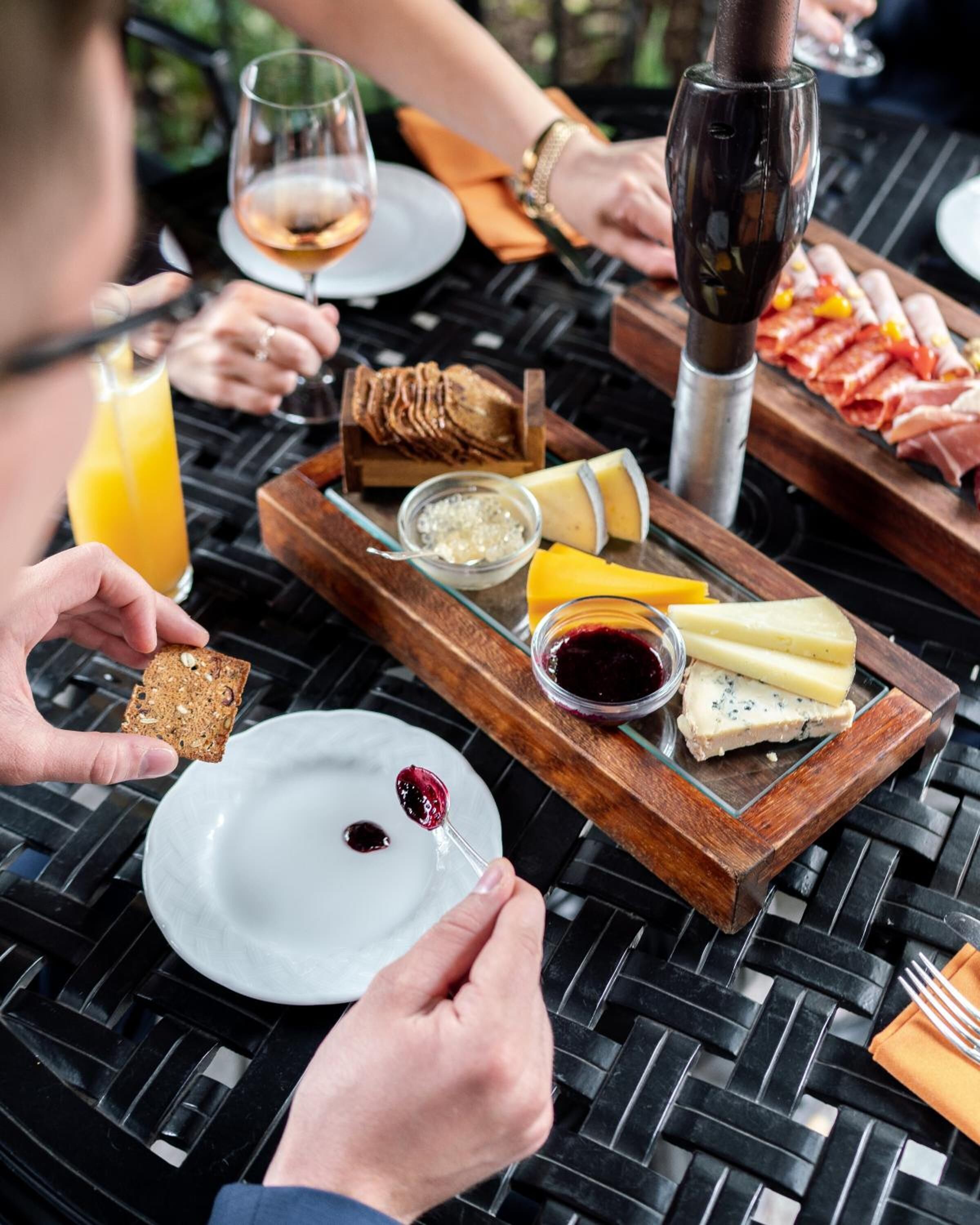 A breakfast setting with a variety of cheeses, bread, and meats on a wooden board, accompanied by a glass of orange juice and a glass of wine.