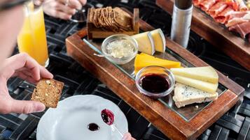 A breakfast setting with a variety of cheeses, bread, and meats on a wooden board, accompanied by a glass of orange juice and a glass of wine.