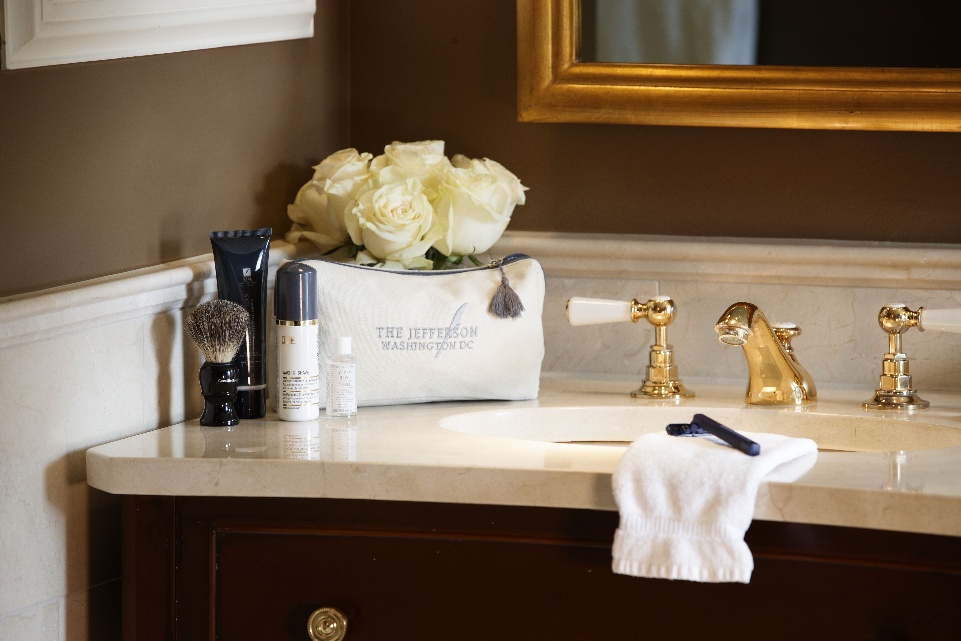 A bathroom vanity with a white marble countertop, gold faucets, a white towel, and a bag of white roses.