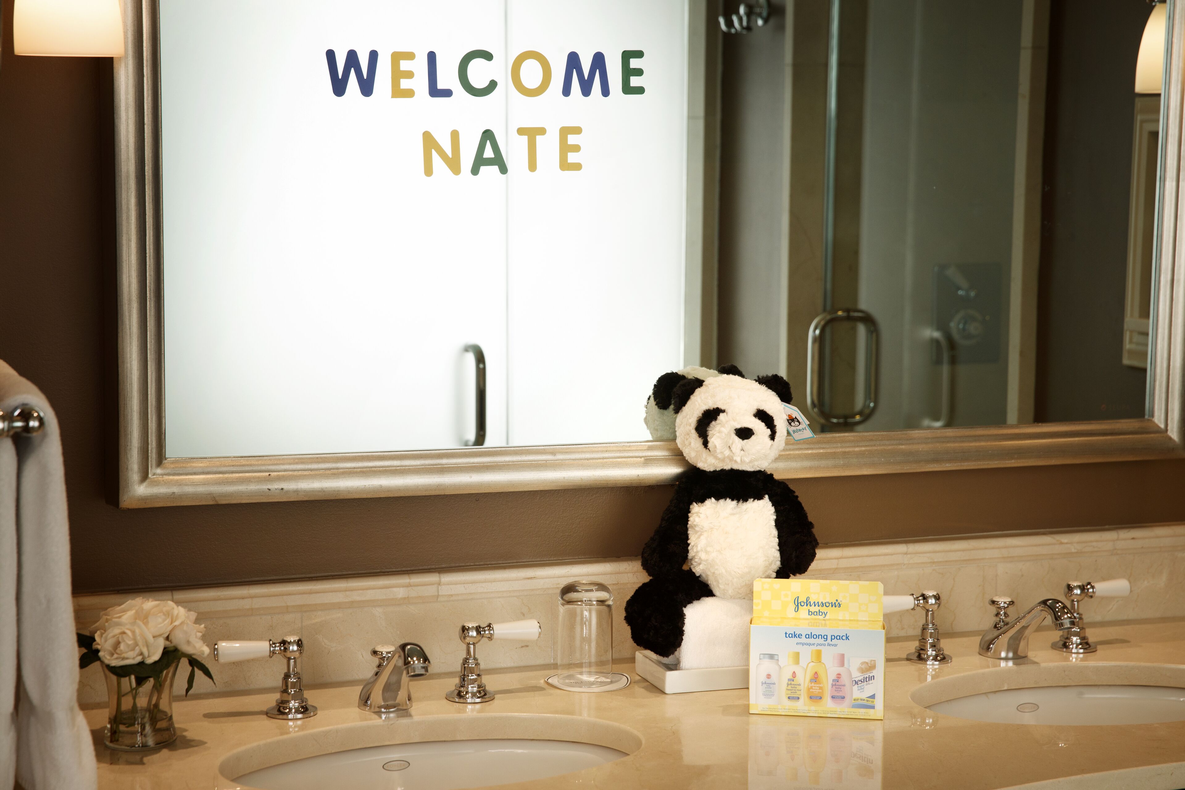 Een badkamer met een spiegel, twee wasbakken, een panda pluchen knuffel en een welkomstbord.