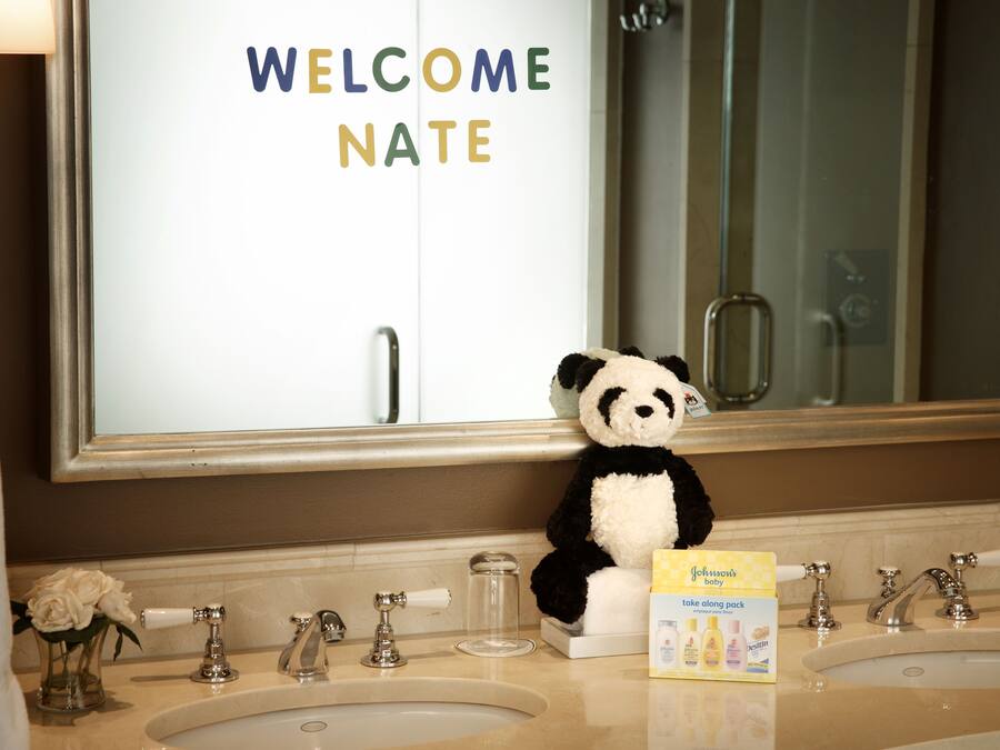 A bathroom with a mirror, two sinks, a panda plush toy, and a welcome sign.