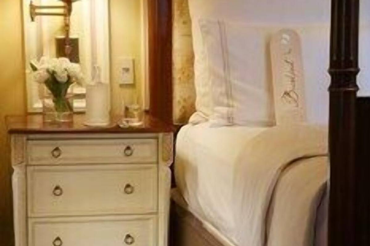 A bedroom with a four-poster bed, a nightstand with drawers, a lamp, and a vase with flowers.