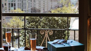 A dining table with plates of food, glasses of wine, and a gift box, viewed through a window overlooking a cityscape with buildings and a tall monument.