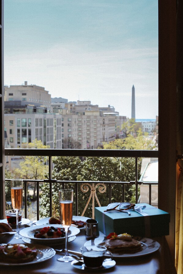 A dining table with plates of food, glasses of wine, and a gift box, viewed through a window overlooking a cityscape with buildings and a tall monument.