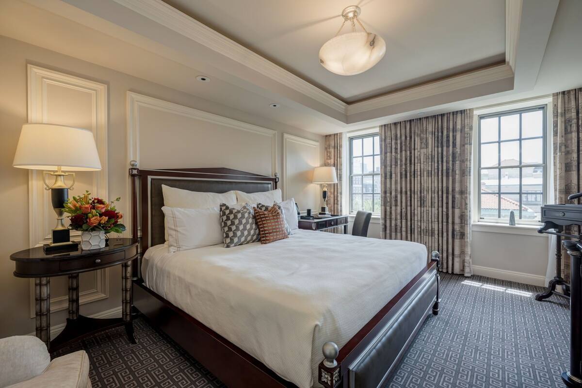 A hotel room with a large bed, two bedside tables with lamps, a desk, and a view of the city through large windows.