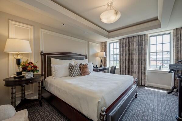 A hotel room with a large bed, two bedside tables with lamps, a desk, and a view of the city through large windows.
