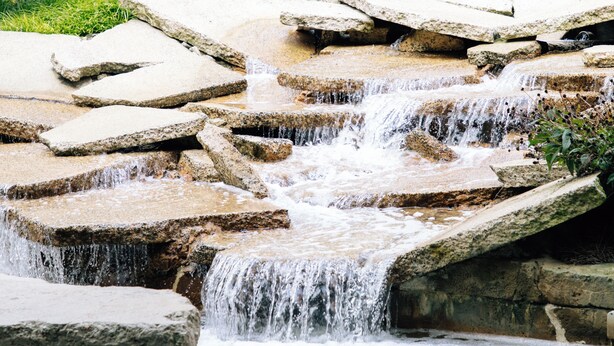 A waterfall cascading over irregularly shaped rocks into a pond, surrounded by lush greenery and a building in the background. - Millennium Hotel Durham (Durham)