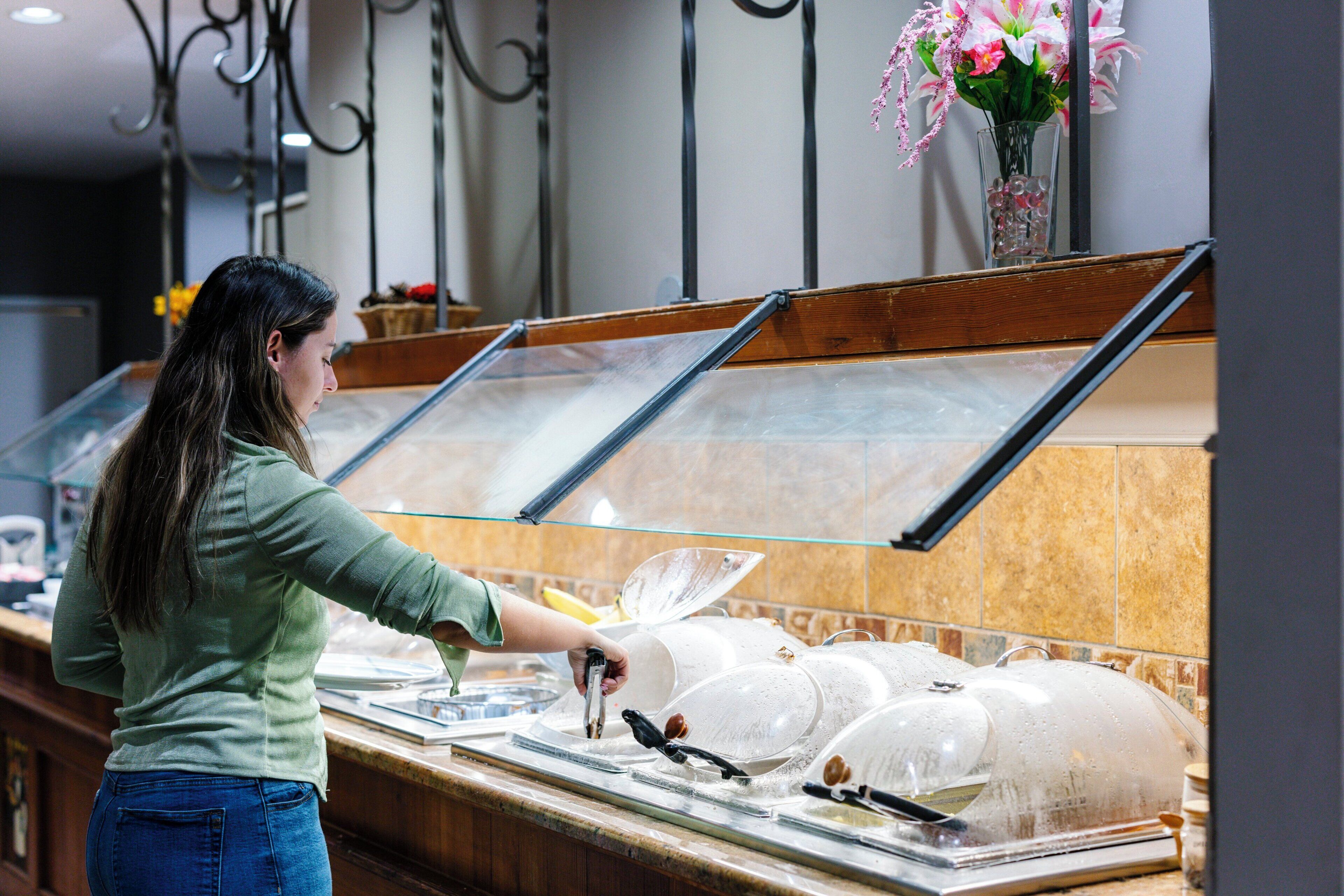 A buffet area with a person serving food, a glass display case, and a vase with flowers on the wall.