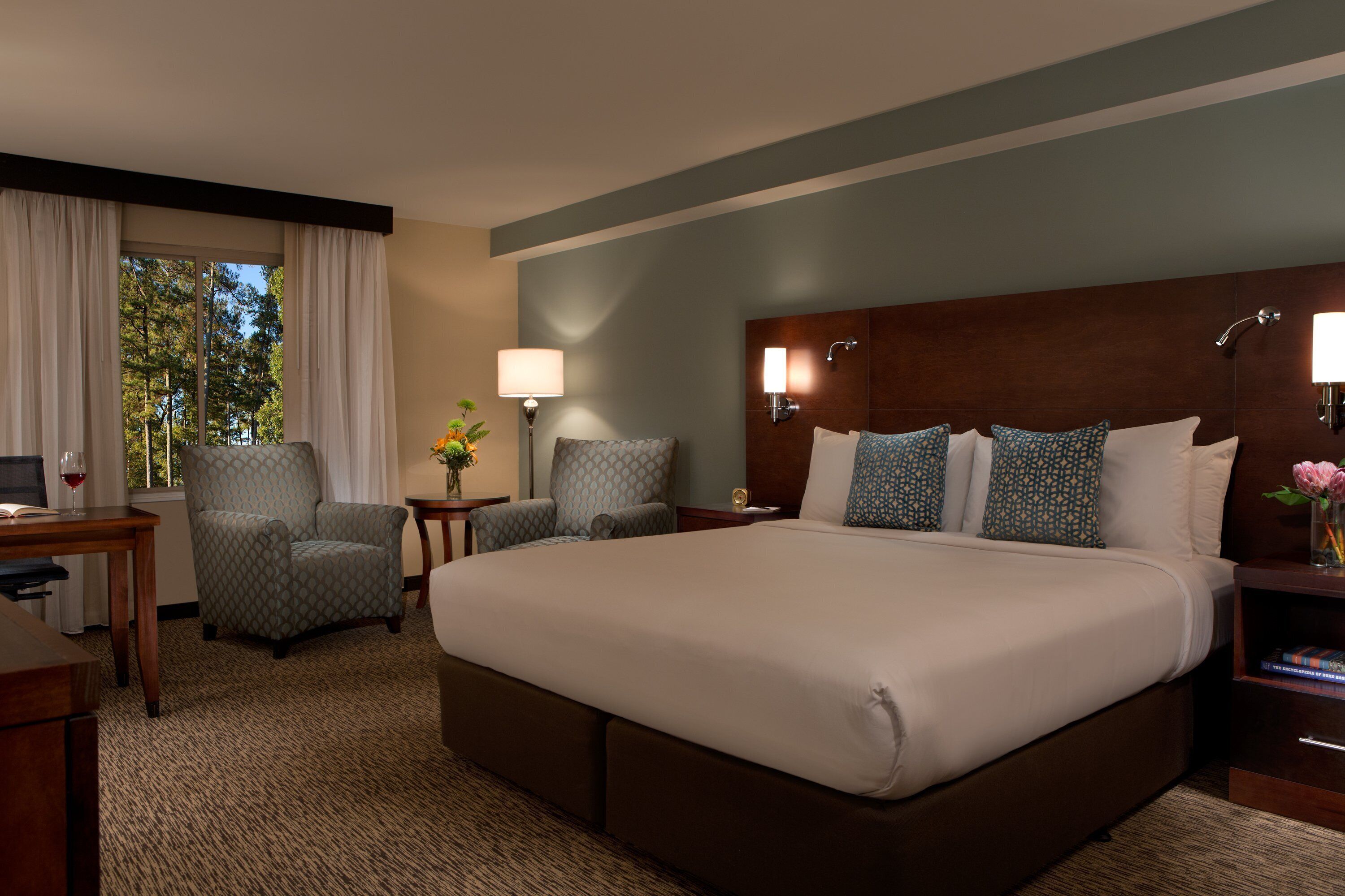 Une chambre d’hôtel avec un grand lit, deux fauteuils, un bureau avec une lampe et une vue sur les arbres par la fenêtre.