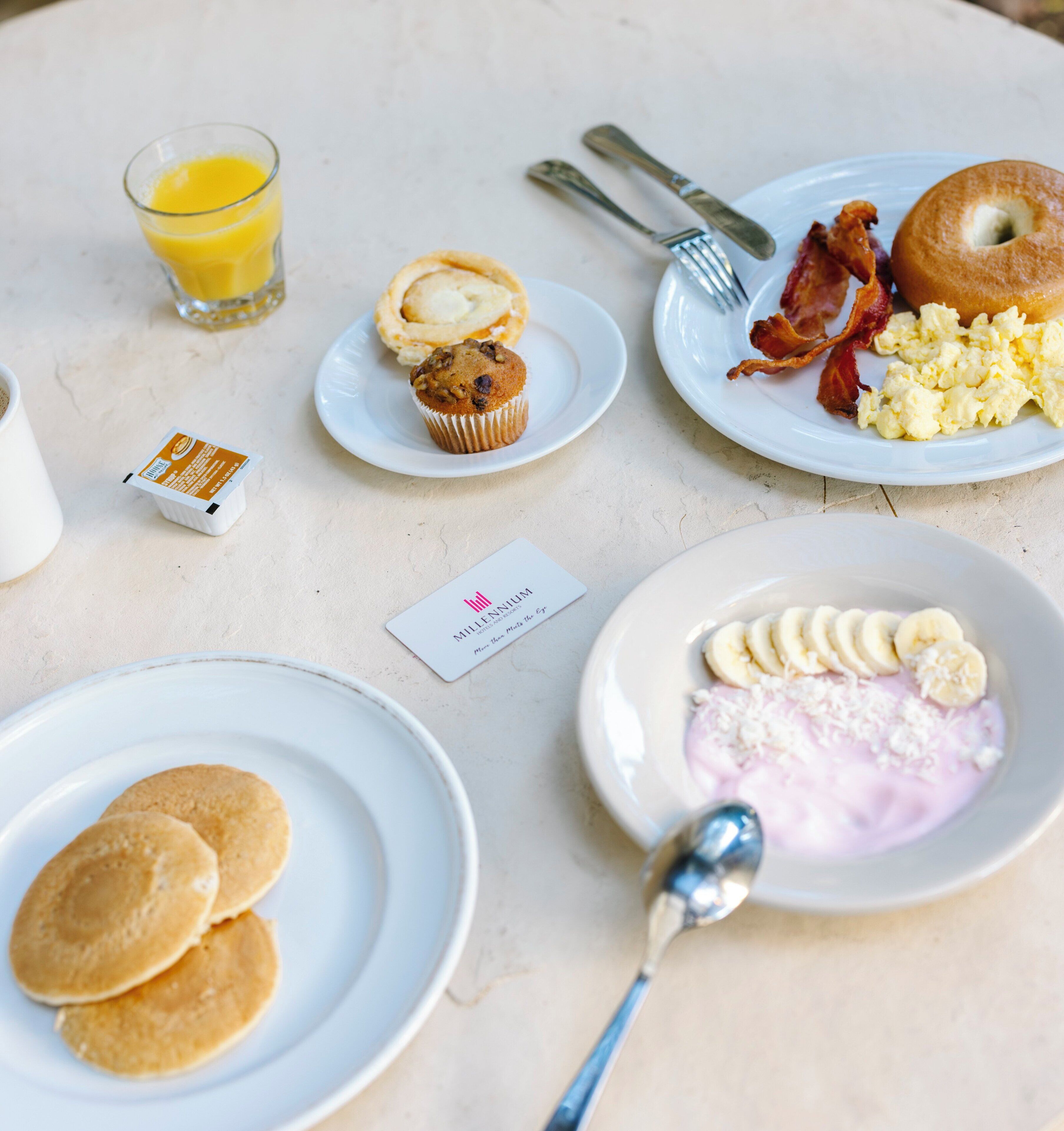 En morgenmadsopsætning med pandekager, muffins, bacon, æg og en smoothie på tallerkener.