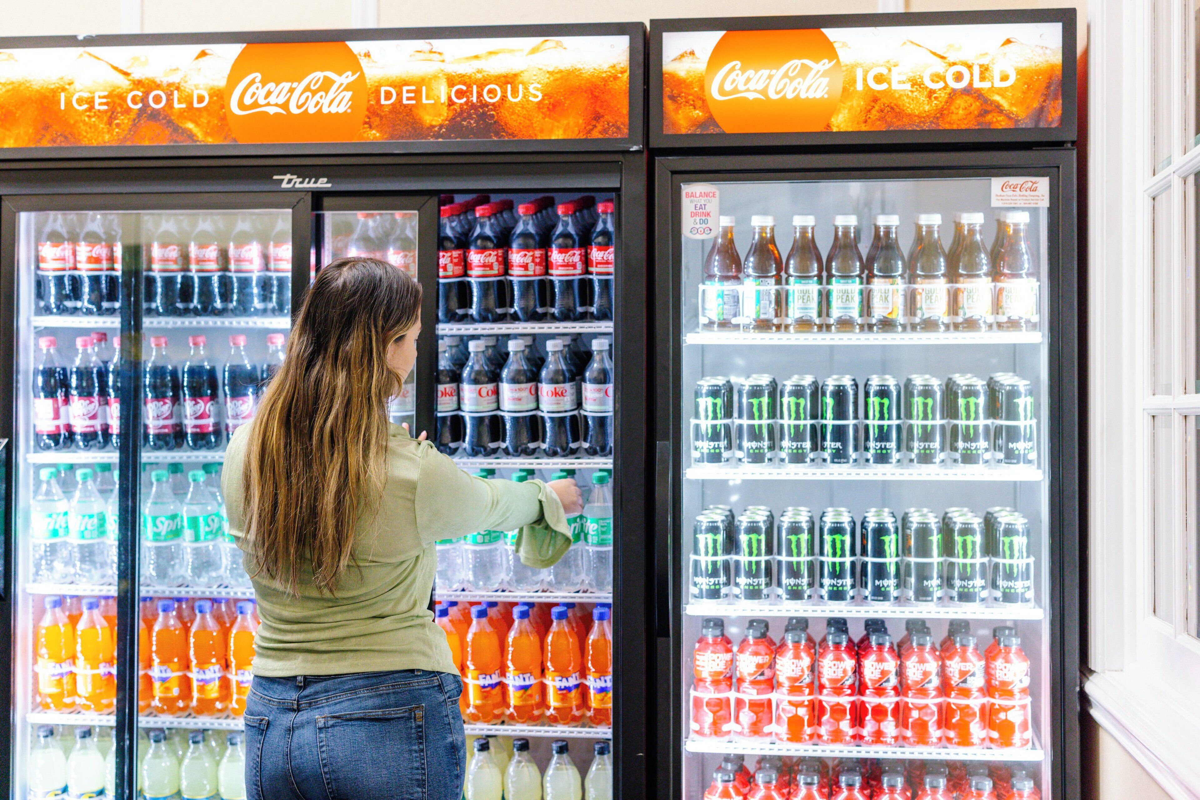 Uma pessoa escolhendo bebidas em uma geladeira Coca-Cola cheia de diversas opções.