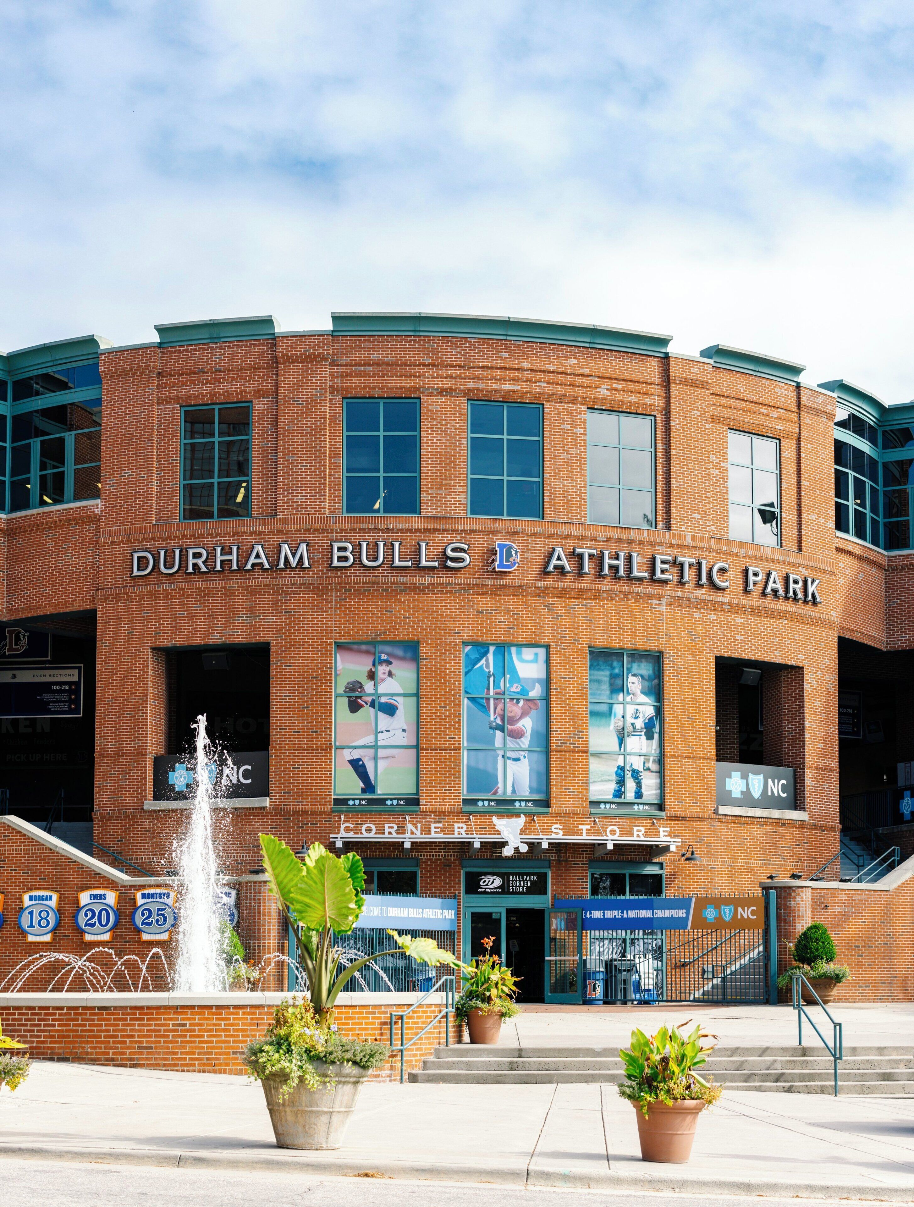 A brick building with 'Durham Bulls Athletic Park' signage, a fountain, and potted plants.