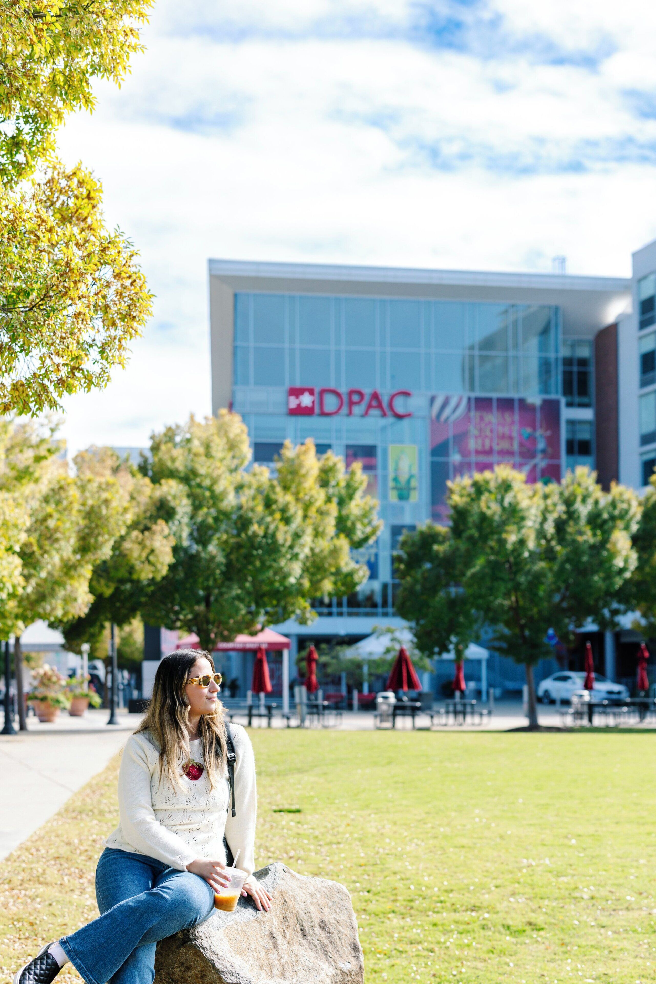 A person sitting on a rock with a drink, a building with 'DPAC' signage in the background, and trees around.