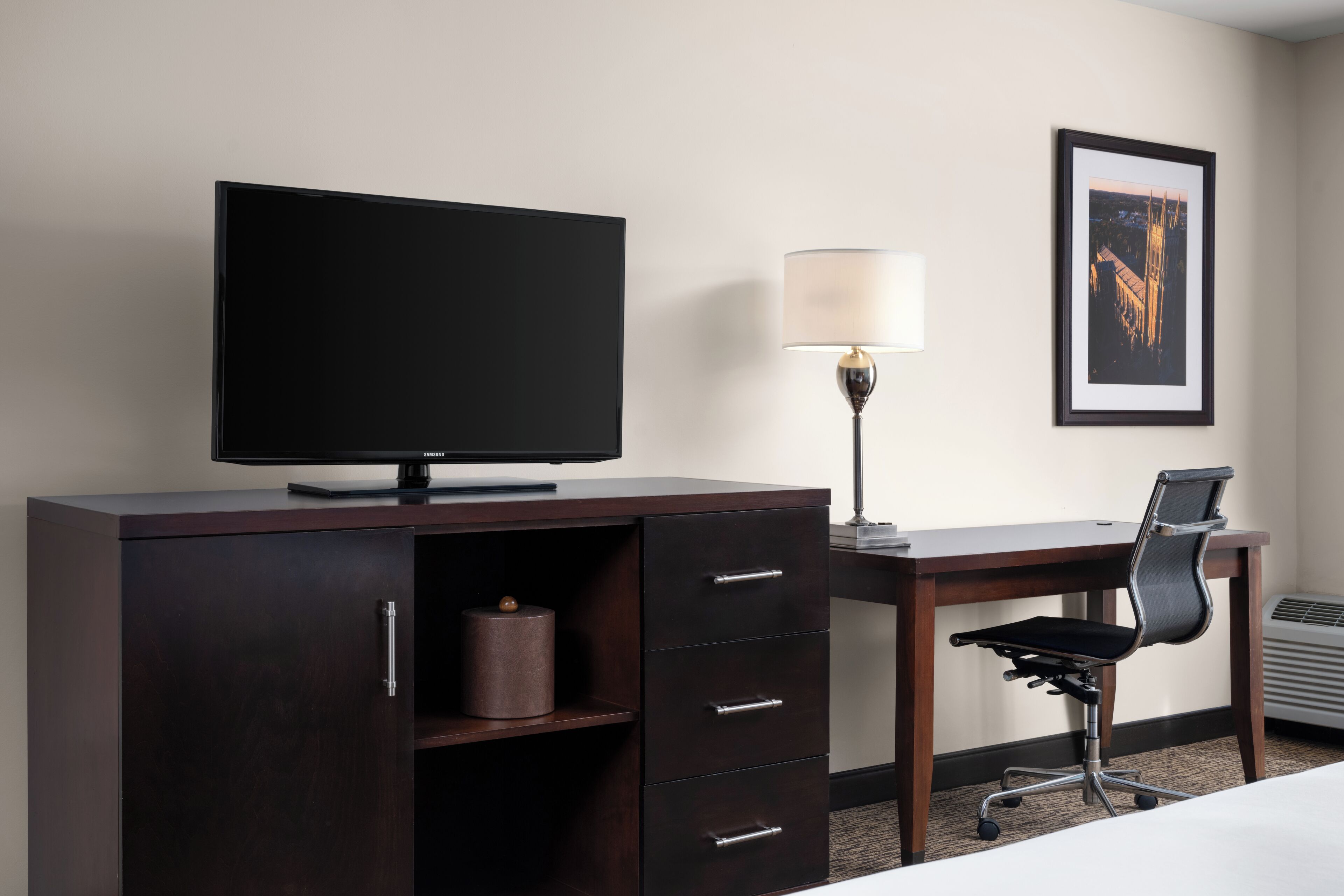 A hotel room with a dark wooden desk, a flat-screen TV, a lamp, a framed picture on the wall, and a black office chair.